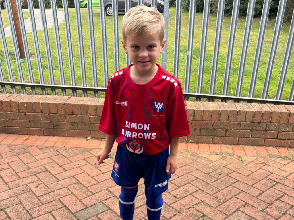A little boy in a red and black football kit is smiling for the camera.
