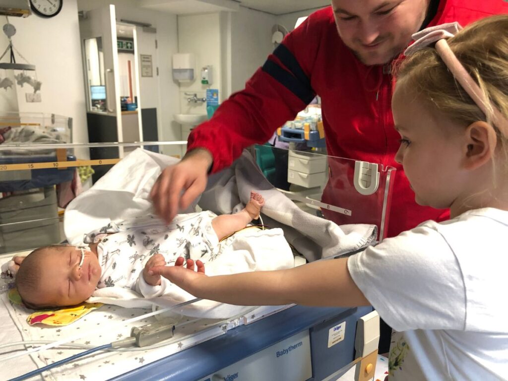 A man and a little girl are talking to a small baby in hospital.