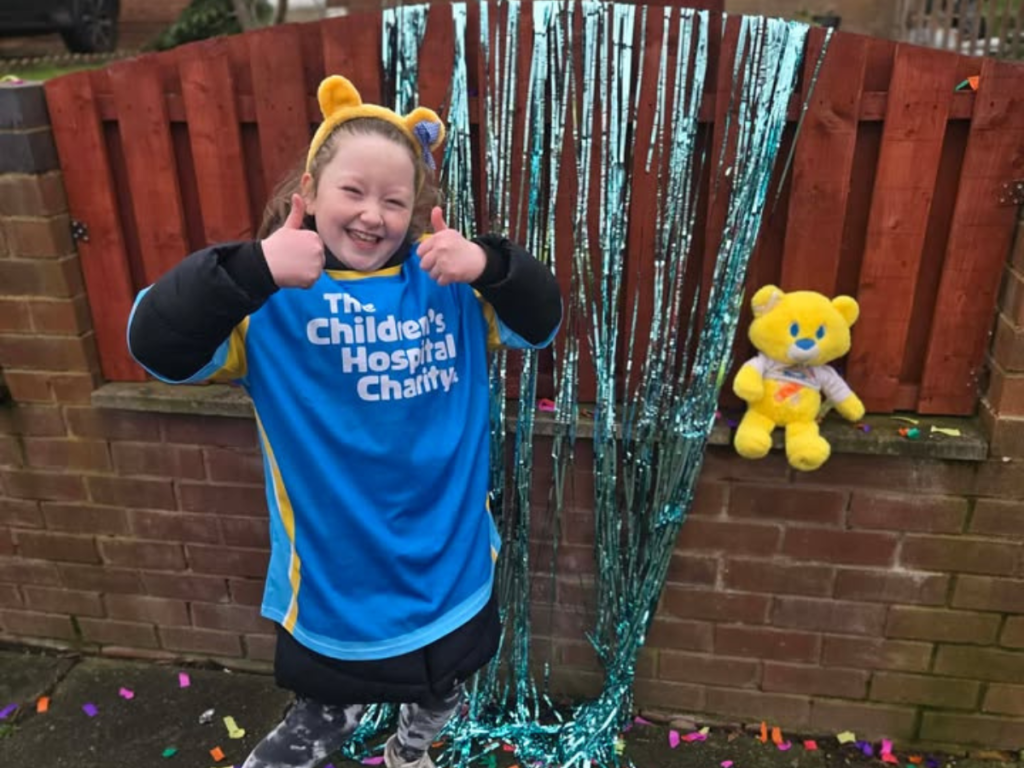 Young girl stands with her thumbs up in front of a glittery backdrop. She wars a charity tshirt and has a Theo bear with her.