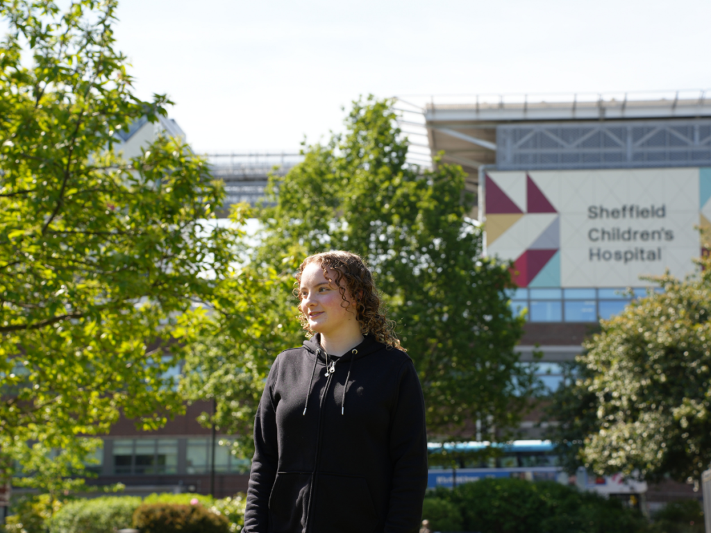A teen girl stands in a park with Sheffield Children's Hospital in the background.