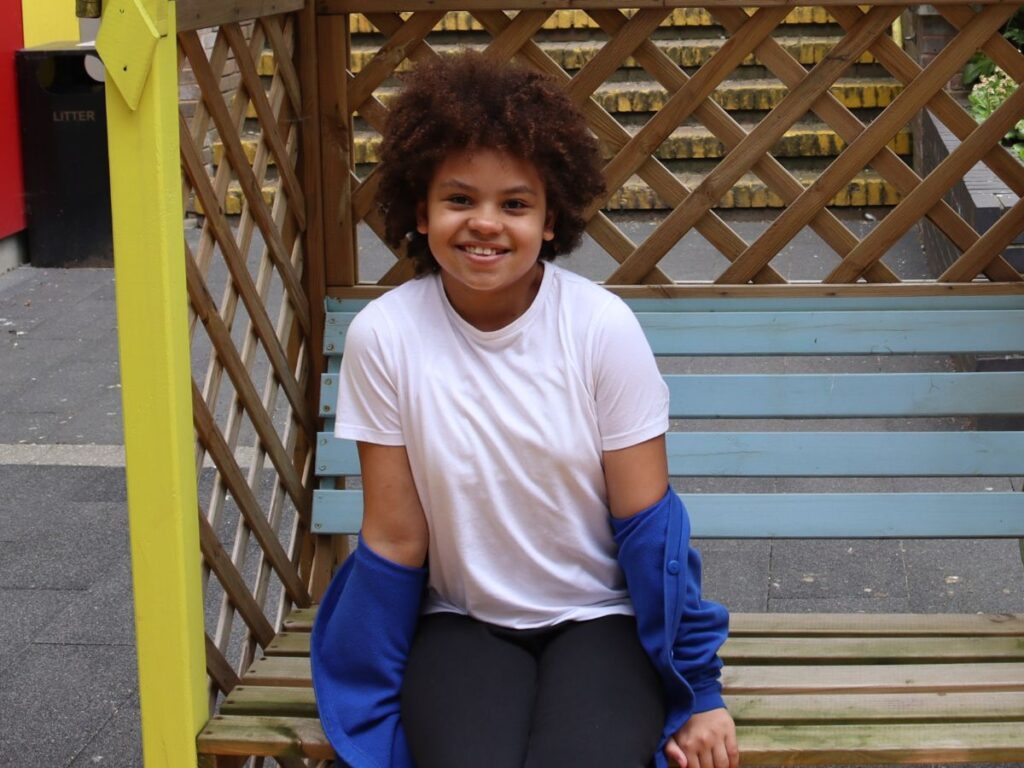 A little girl is sitting on a bench wearing a white polo shirt and a blue cardigan. She is smiling for the camera.