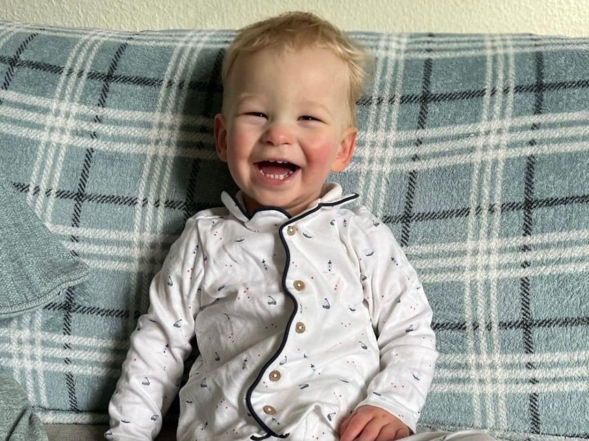 A young boy in pyjamas with sail boats on them is sitting on a tartan sofa and laughing.