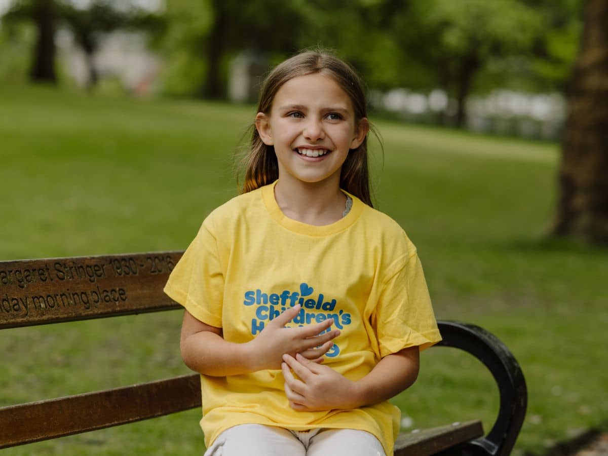 A young girl sits on a park bench. She is wearing a yellow Sheffield Children's Hospital Charity t-shirt.