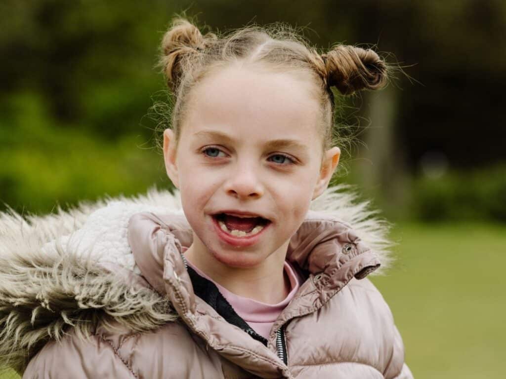 A young girl is standing in a park and smiling. She has a cosy coat on and her hair is tied in bunches.
