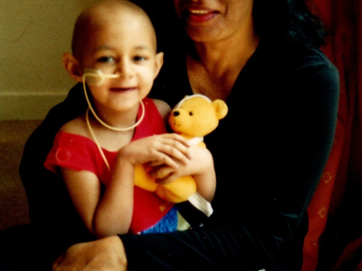 An old photograph of a young girl in a red t-shirt holding a yellow teddy bear. She has an NG tube in her nose.