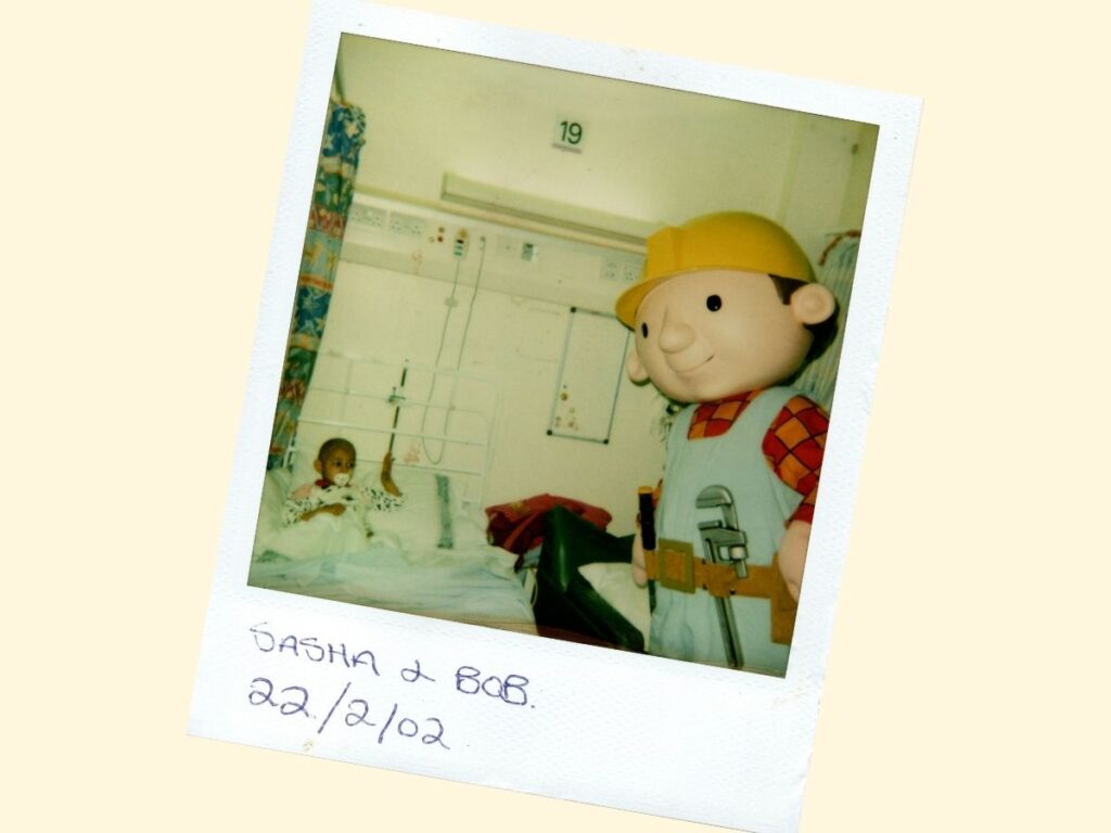 A young girl in a hospital bed is visited by a large mascot character dressed as a builder.
