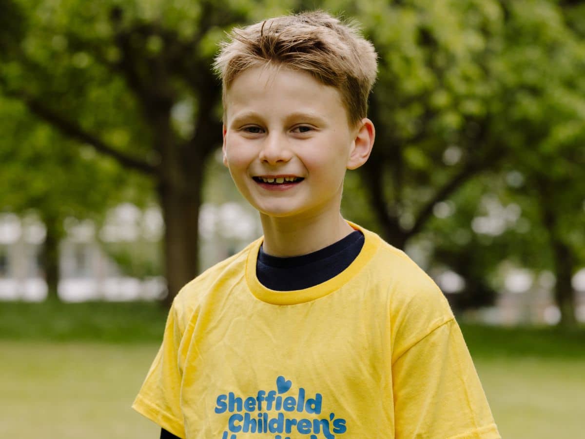 A young lad is standing in a park smiling. He is wearing a charity-branded yellow t-shirt with blue writing on it.