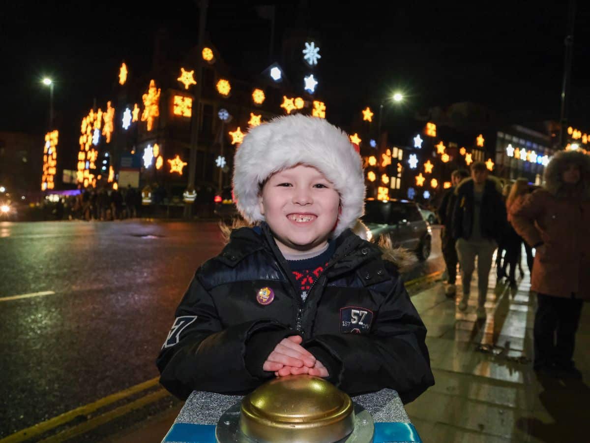 A young boy wearing a Santa hat is pressing a large button with a large snowflake light display on a building behind him.