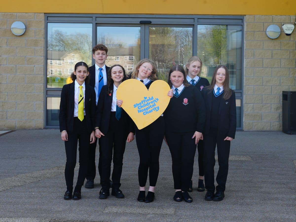 A group of teenagers are standing outside a colourful building holding a big yellow heart with a blue charity logo on it.