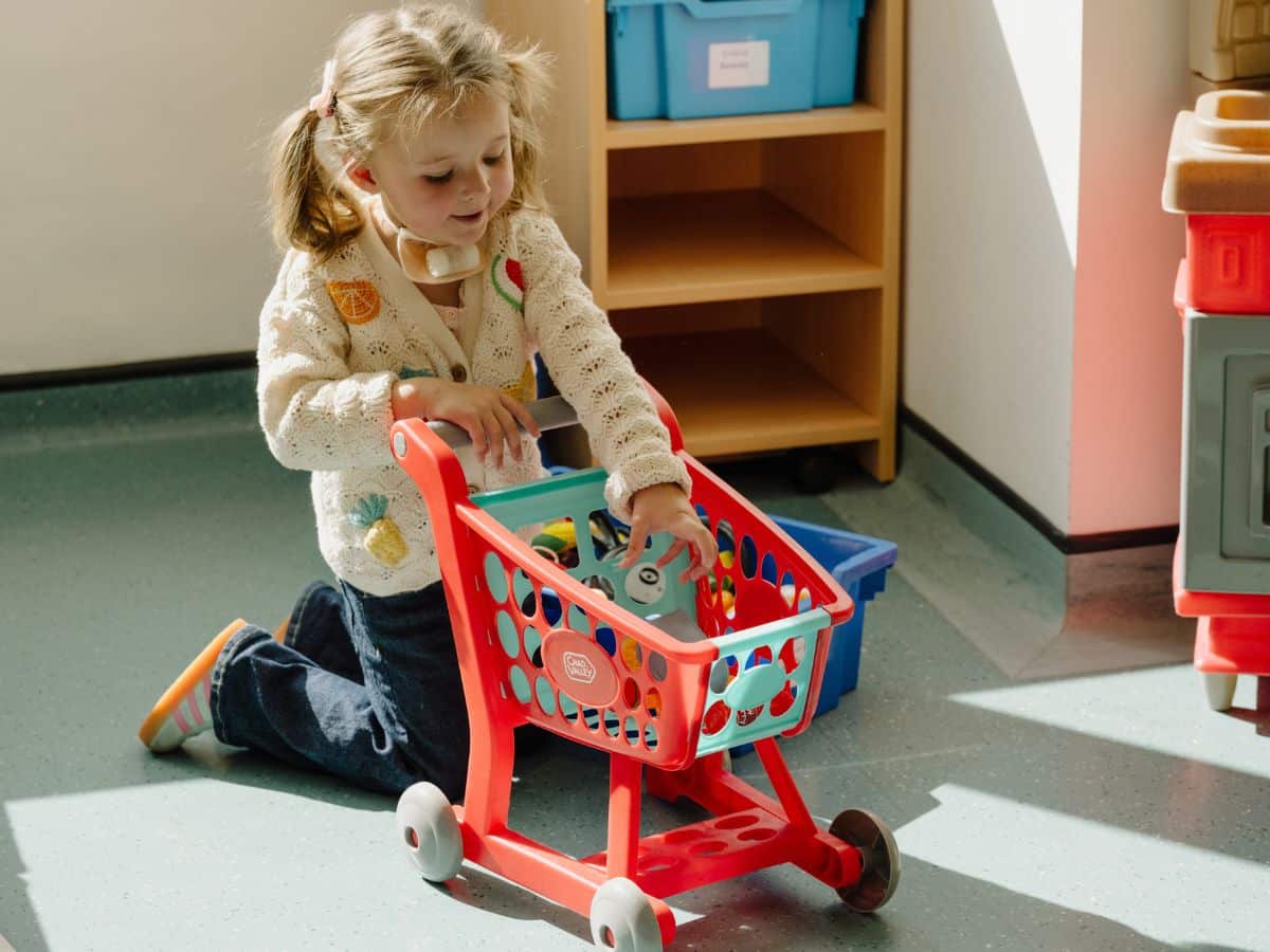 A little girl with dressing on her throat is playing with a toy trolley full of make-believe grocery items.