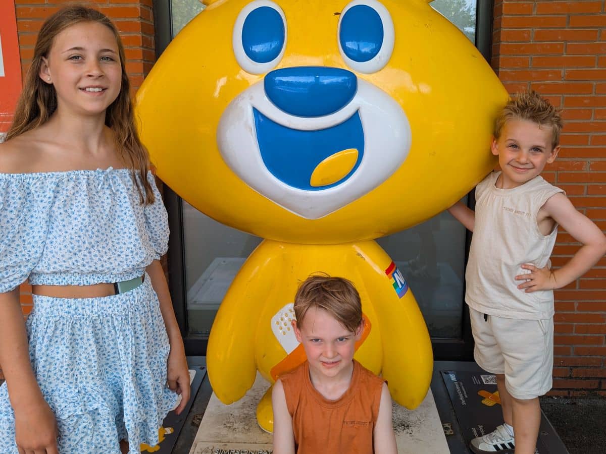 Three children stand next to a statue of a yellow bear.