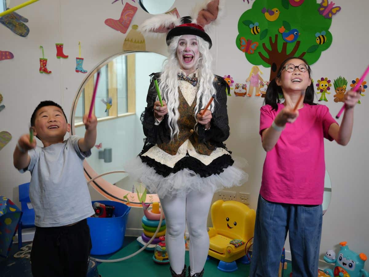 A lady dressed as a white rabbit is doing a circus skills workshop with two small children in a colourful room.